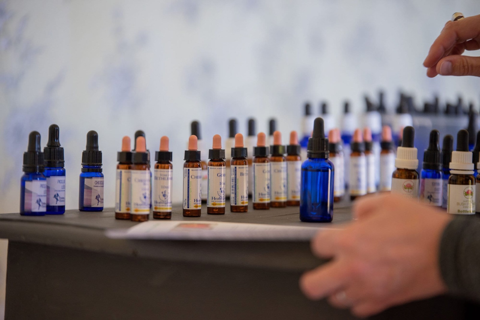 Dozens of blue and brown dropper bottles of flower essences laid out on a dark table in the garden room, with Lucy's hands in frame