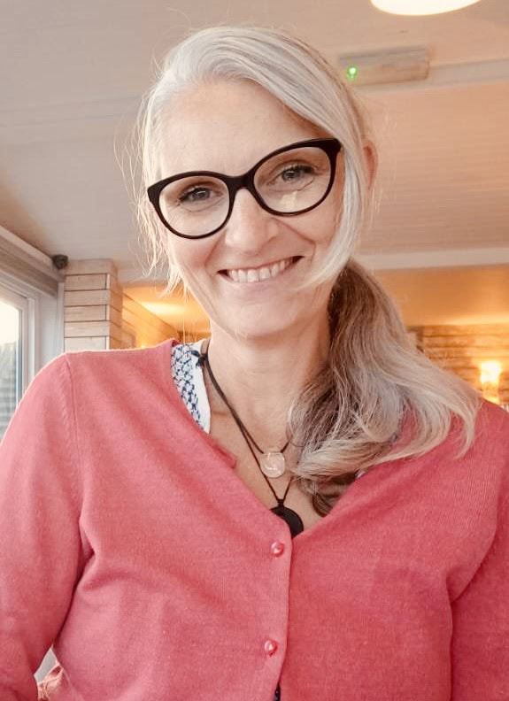 Lucy Hunter, smiling, in glasses and a coral cardigan, in her garden room